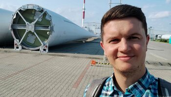Zdjęcie przedstawia absolwenta UPWr, kierunku Inżynieria Bezpieczeństwa - Marcina Robaka. W tle, na placu widać łopatę turbiny wiatrowej.