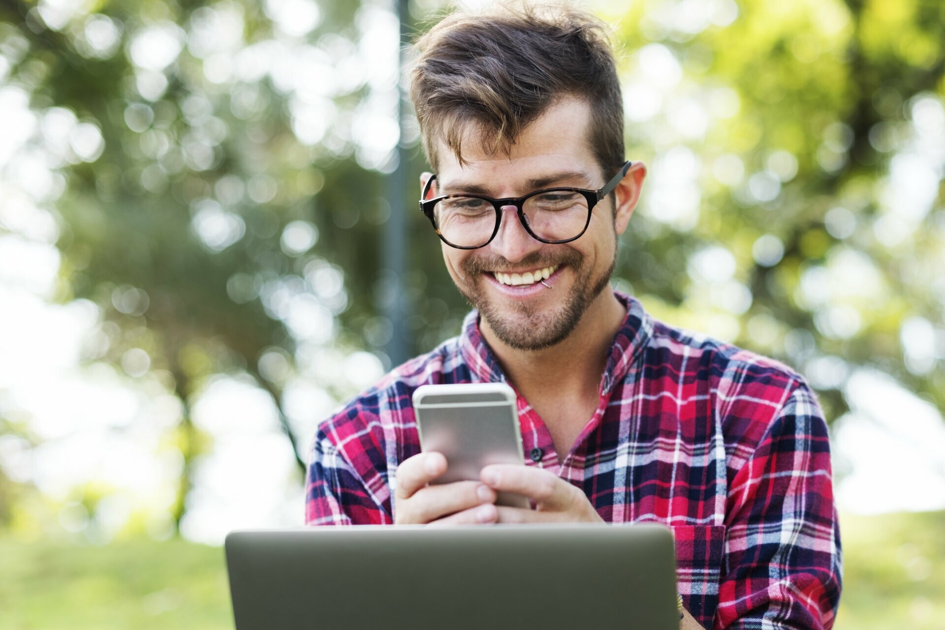 Na zdjęciu znajduje się uśmiechnięty student w okularach, trzymający smartfona. Przed nim widać też ekran laptopa.