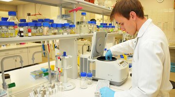 Na zdjęciu znajduje się mężczyzna w laboratorium chemicznym, umieszczający materiał do badań w urządzeniu. Wokół niego na blacie i półkach znajduje się dużo naczyń z materiałami chemicznymi.